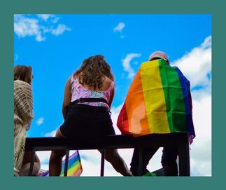 back of three people sitting on a bench one has the rainbow flag over their shoulder